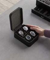 A hand reaching towards the TAWBURY Fraser 4 Watch Travel Case - Black from the Fraser Collection, containing four wristwatches, displayed on a gray surface.